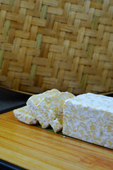 A block of tempeh, partially sliced, sits on a wooden cutting board with a woven bamboo mat in the background.