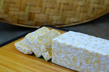 A block of tempeh, partially sliced, sits on a wooden cutting board with a woven bamboo mat in the background.