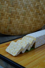 A block of tempeh, partially sliced, sits on a wooden cutting board with a woven bamboo mat in the background.