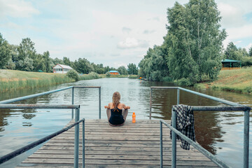 woman sitting cross-legged on wooden dock over calm river, wearing sportswear, back view, summer greenery and countryside houses, meditation, morning ritual, nature therapy