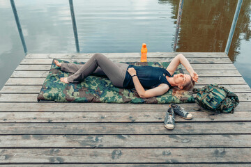 person lying flat on pier with bottle and jacket near, lake reflection and tree branches above,...