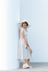 Woman wearing a white polka dot dress and straw hat stands against a smooth white column, holding a beaded handle handbag. Architectural lines and soft summer shadows in a minimalist outdoor scene