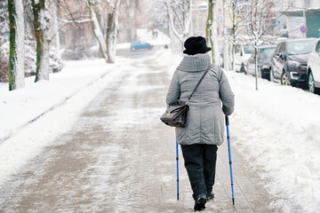 Senior woman walking with canes for stability on hazardous snow covered and icy sidewalk during frosty winter day. Woman walking with nordic poles