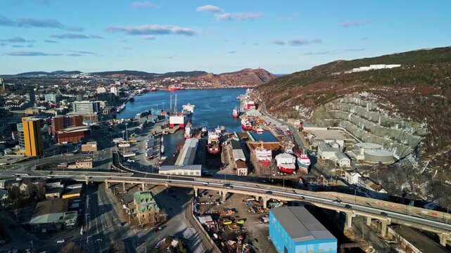 A drone glides above St. John's, Newfoundland, capturing an elevated highway along the harbour, downtown buildings, ships moored at the waterfront, and hillsides meeting the Atlantic.