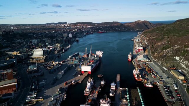 A sweeping drone shot looks down the length of St. John's harbour in Newfoundland, showing downtown buildings on one side, ships and docks on the other, and hills rising above the Atlantic.