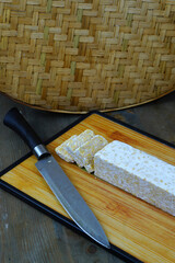 A block of tempeh, partially sliced, on a wooden cutting board with a knife, set against a woven bamboo background.