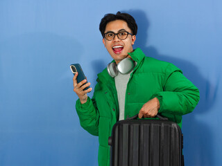 Young Asian man in green winter jacket looking amazed while holding a phone and suitcase, ideal for winter holiday ads and promotions.
