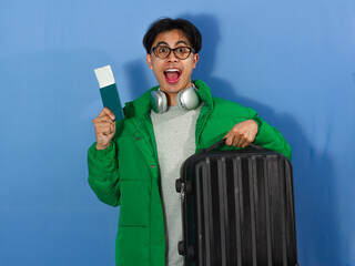 Excited young Asian man in a green winter jacket holding a passport and suitcase, surprised...