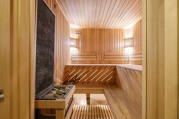 well-lit, wooden sauna featuring benches, a stone heater, and warm lighting. The room exudes a...