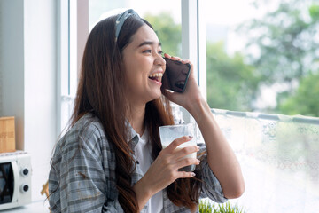 Beautiful Asian girl talking happily on the phone while holding a glass of milk she drinks in the morning in her other hand.