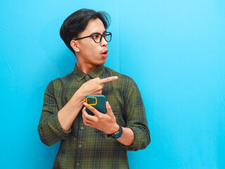 Attractive Asian man holding mobile phone showing shocked expression isolated on blue background
