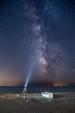 Man standing on a beach next to an abandoned boat, pointing a bright light beam into the Milky Way galaxy. Vertical astrophotography of a starry night sky over the sea. Concept of cosmic journey.