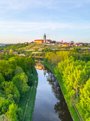 Aerial view showcases Melnik City and its historic chateau perched above the lush banks of the Labe River. The scene captures the vibrant greenery and tranquil waters at sunset.