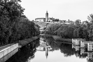 Chateau Melnik stands majestically above the calm waters, its reflection creating a mirror-like effect at twilight. Lush trees and sloping hills surround the serene landscape.