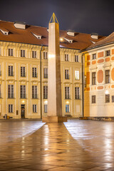 The Mrakotin Monolith stands tall at Prague Castle, surrounded by historical buildings, as warm light casts dramatic shadows on the cobblestone ground under the night sky.