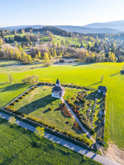Located in Korenov, the Tesarov Chapel is framed by a quaint graveyard, surrounded by lush greenery and rolling hills of the Jizera Mountains, capturing a peaceful, serene atmosphere.