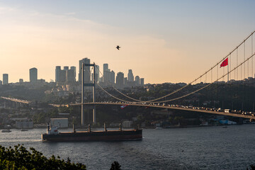 Obraz premium Bosphorus Bridge in Istanbul during golden hour with a large cargo ship passing underneath. Modern city skyline and skyscrapers in the background. Concept of international trade and Turkish tourism.