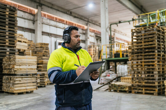 Supervisor wearing ear protection and high visibility jacket writing notes on clipboard while inspecting wooden pallets in a recycling factory