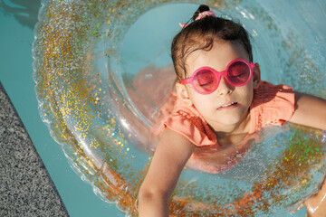 Cute funny toddler girl in a sunglasses relaxing on inflatable toy ring floating in pool have fun...