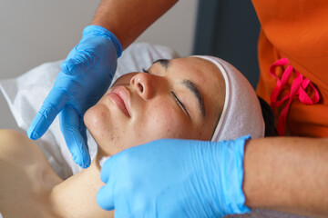 Beautician wearing blue gloves applying anti acne treatment to young woman's face lying on a white towel