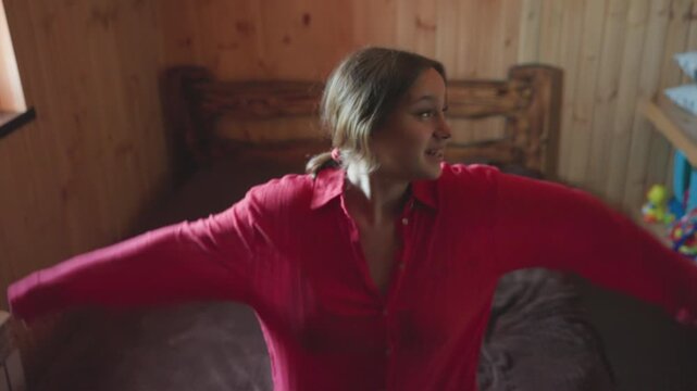 lady in red top smiles joyfully while sitting on bed in wooden room then gently falls backward on bed playfully showing carefree moment filled with happiness in cozy indoor rustic setting