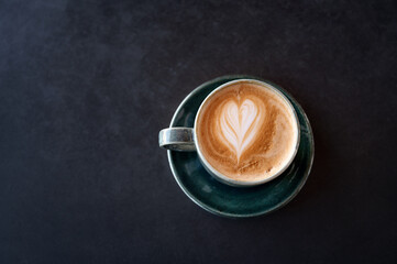 Flat lay of a green ceramic cup filled with latte, showcasing heart-shaped latte art. The cup sits on a dark textured surface, creating a minimalist and modern coffee shop aesthetic.