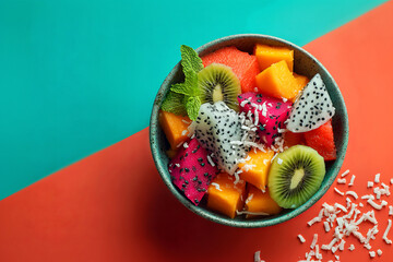 Healthy fruit salad bowl with exotic fruits like pitaya, papaya, and mint leaves on turquoise and coral backdrop.