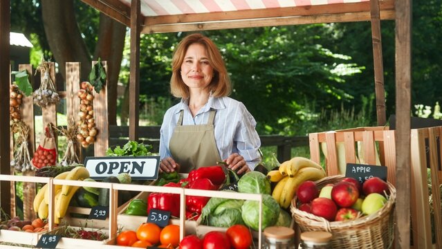 Positive Caucasian female farmer standing at open stall and waiting for customers. Cute woman cheerfully smiling and selling her fresh, natural vegetables and fruits. Agriculture concept.