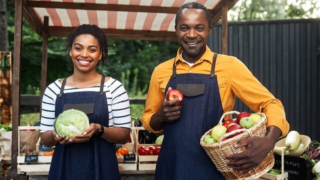 Happily African American couple sellers with black aprons standing in front of food market. Positive husband holding basket full of fresh apples while pretty wife with cabbage smiling on camera.