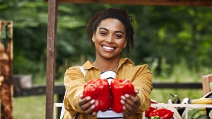 Charming African American woman showing on camera organic red peppers at foods market. Positive...