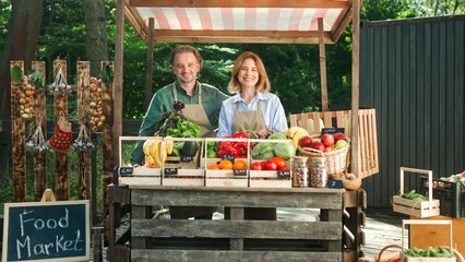 Hardworking Caucasian couple farmers with aprons standing at local food market. Positive family getting ready for selling fresh vegetables, herbs and fruits. Working together. Farming concept.