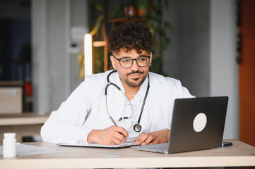 Arab male doctor working on laptop and taking notes in modern clinic