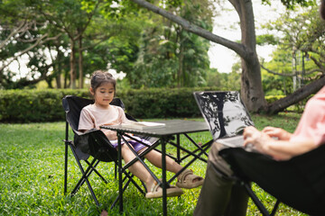 Young girl sitting alone at a table in a garden, while an adult works on a laptop in a peaceful outdoor environment with greenery