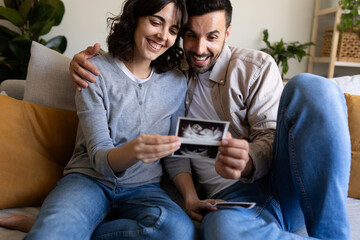Happy pregnant couple looking baby ultrasounds at home sitting on the couch