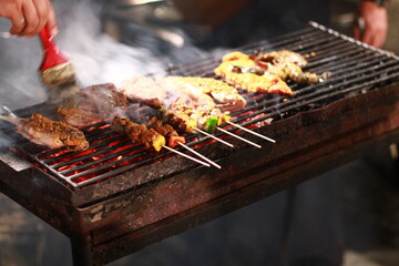 
A person uses a basting brush to coat various meats, including fish, skewers, and other cuts, on a...