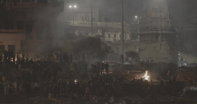 Varanasi, Uttar Pradesh, India. Cremation in process on Manikarnika Ghat. Cinematic Camera movement moving along riverbank embankment. View from riverside. night lights. Fire, smoke - Funeral rites of