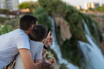Father and Son Enjoying a Moment at a Waterfall in a Scenic Park During Golden Hour, Capturing the...