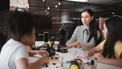 White female teacher advises and instructs primary girls group students, Robotic arms design at learning table in science classroom, discusses experiment knowledge for children of elementary school.