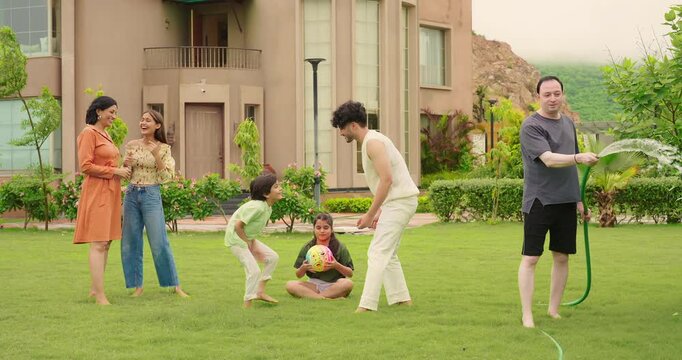 Indian happy dad man hold hose pipe spray water on family group outdoor house lawn park young adult teen girl mom cute son kid boy child male female play game have fun joy spend time enjoy picnic trip