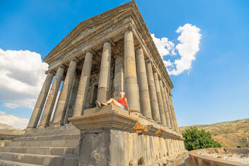 Blonde tourist resting on the steps of the Garni Temple, a hellenistic temple located in the village of Garni, Armenia, enjoying summer vacation under a blue sky with some clouds