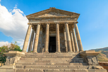 Garni Temple, a well-preserved Greco-Roman colonnaded building, stands majestically on a green lawn, with a bright blue sky and white clouds creating a picturesque backdrop in Garni, Armenia