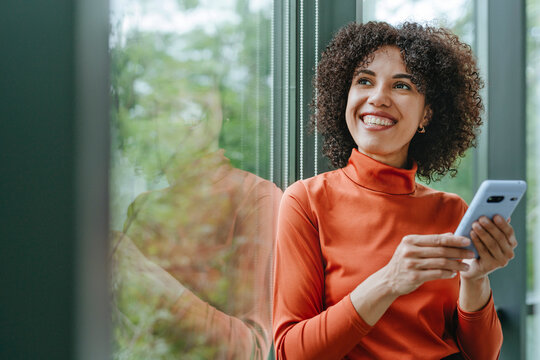 Businesswoman in orange turtleneck using phone and smiling by window - Powered by Adobe