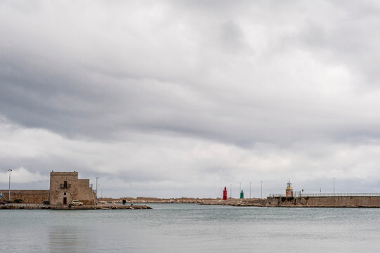 Il cielo coperto di nuvole incontra le acque tranquille di un porto a Trani, in Puglia. Un edificio in pietra si erge lungo il lato sinistro del porto, accanto a una piccolo  faro.