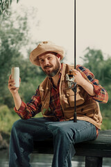 Bearded man in fishing outfit holding beer can and rod while smiling near water. Concept of leisure...