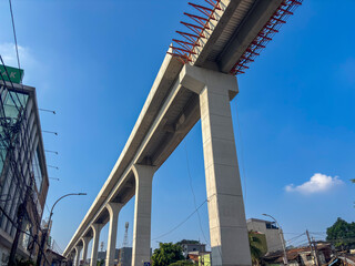 An elevated concrete LRT Jakarta track, under construction, rises on massive pillars against a clear blue sky. Visible red rebar framework highlights ongoing urban infrastructure development and progr