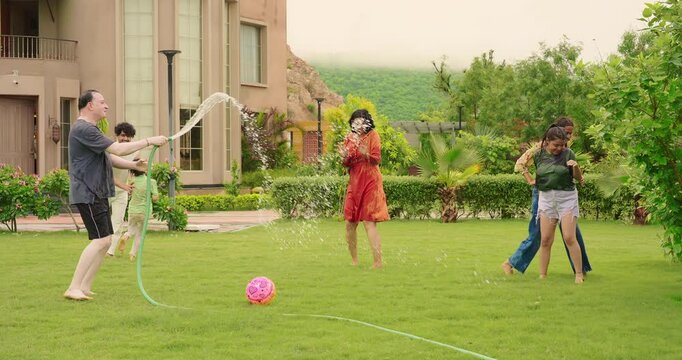 Indian happy dad man hold hose pipe spray water on family group outdoor house lawn park young adult teen girl mom cute son kid boy child male female play game have fun joy spend time enjoy picnic trip