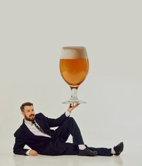 Man in black suit lying on floor balancing oversized beer glass with amused expression. Concept of...