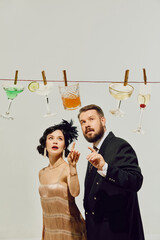 Man and woman in retro party outfits pointing at cocktails clipped to string with surprised faces....