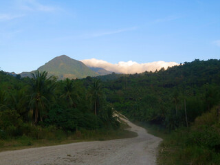 Dirt road in the depths of a tropical island. View of the dirt road with mountains in the background. The dirt road goes through hilly terrain steeply covered with trees.