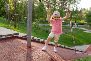 Happy little girl in pink clothes climbed a steep slope holding on to a rope. Physical education at school or kindergarten. Fun sports game on the playground.